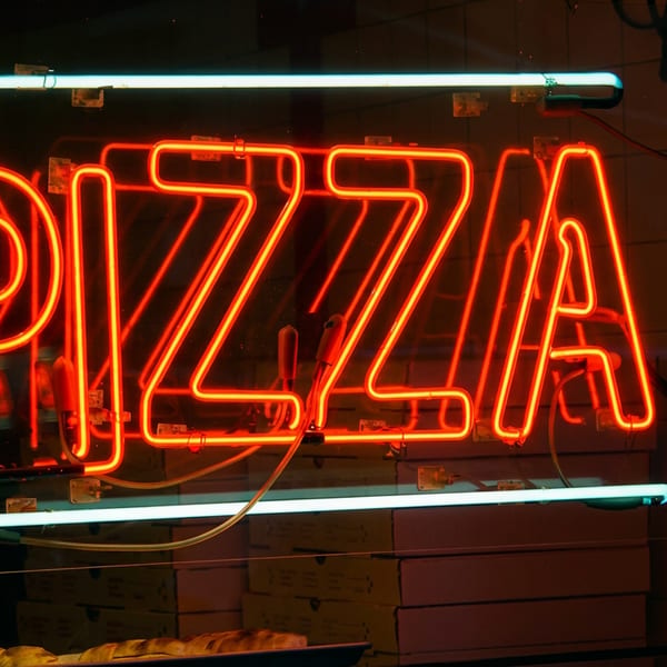 Glowing red and blue pizza neon sign above stacked pizza boxes, with a chef figurine and pizza partially visible below.