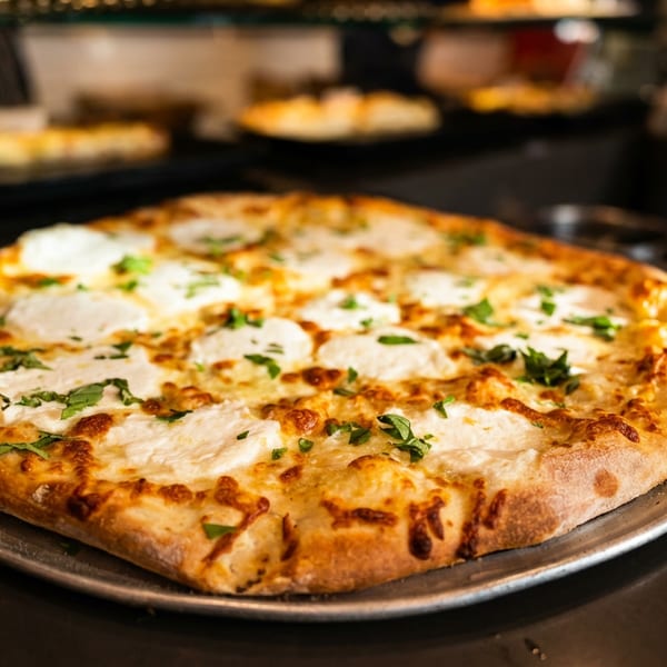 Cheesy, herb-topped pizza with a golden crust sits on a metal serving tray in a restaurant kitchen.