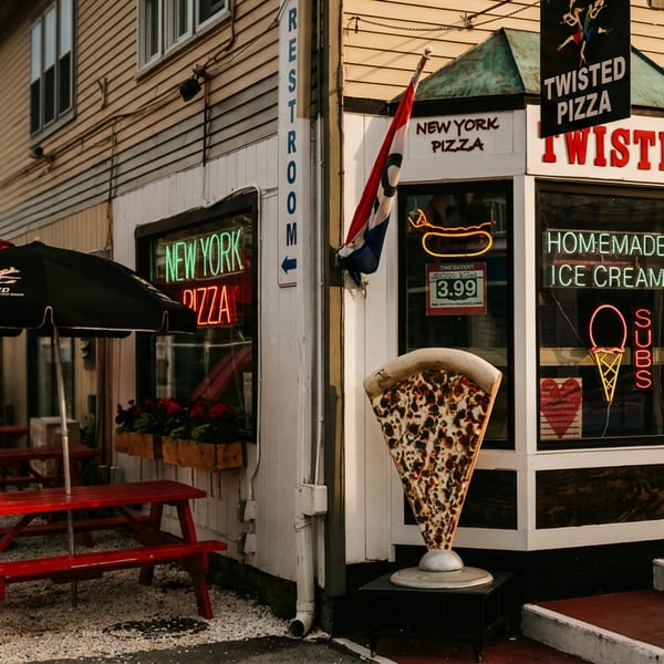 Twisted Pizza storefront with red picnic tables, neon signs, oversized pizza slice, and giant soft-serve cone displays.