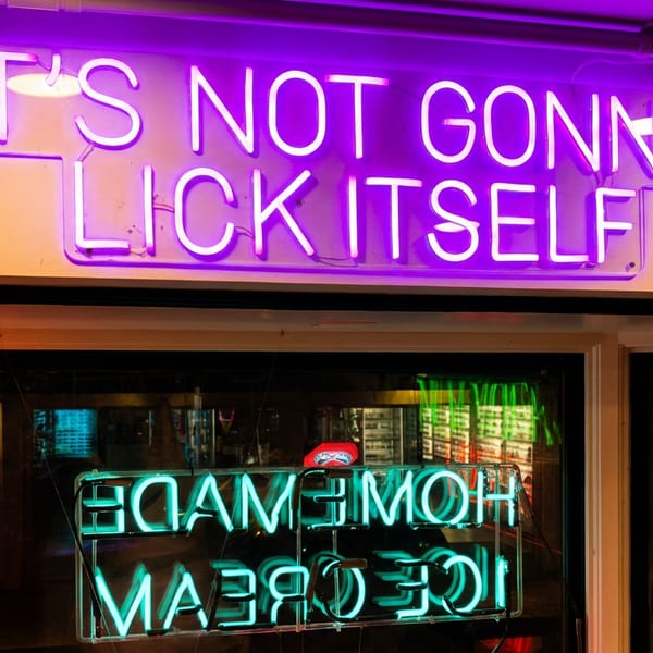 Purple neon sign reading “It’s not gonna lick itself” above a restaurant window with a reversed “Homegrown Ice Cream” reflection.