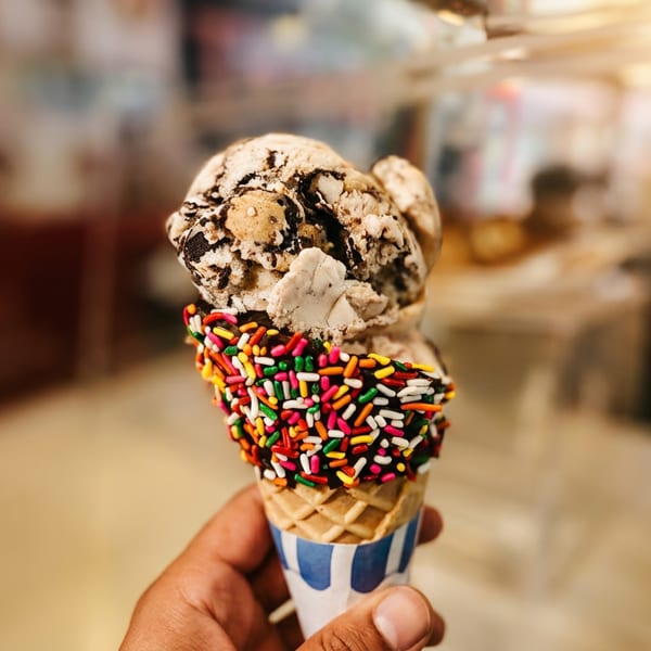 Hand holding a waffle cone with colorful sprinkles, chocolate ice cream, and a cookies-and-cream scoop inside a shop.
