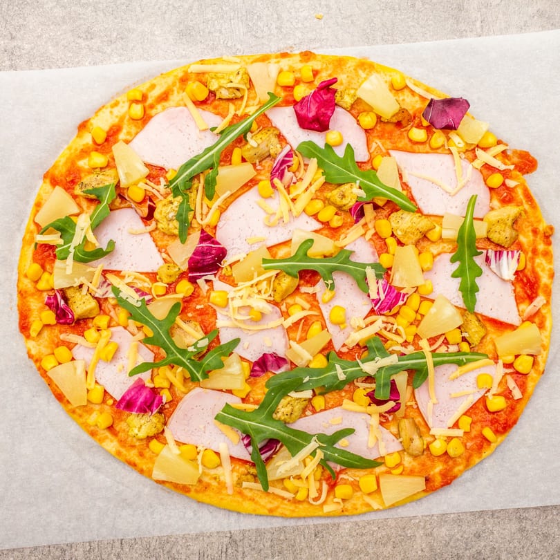 Uncooked Twisted Pizza with ham, pineapple, corn, chicken, cheese, and arugula on parchment paper.